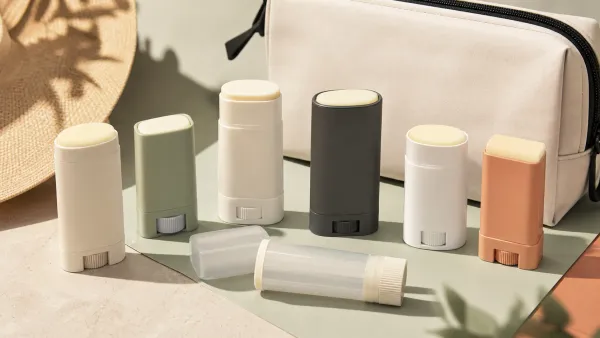Seven unbranded sunscreen sticks arranged with a travel pouch and sun hat on an ivory studio background