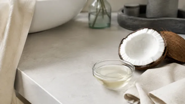 Open coconut oil jar beside fresh coconut halves and green leaves on warm stone
